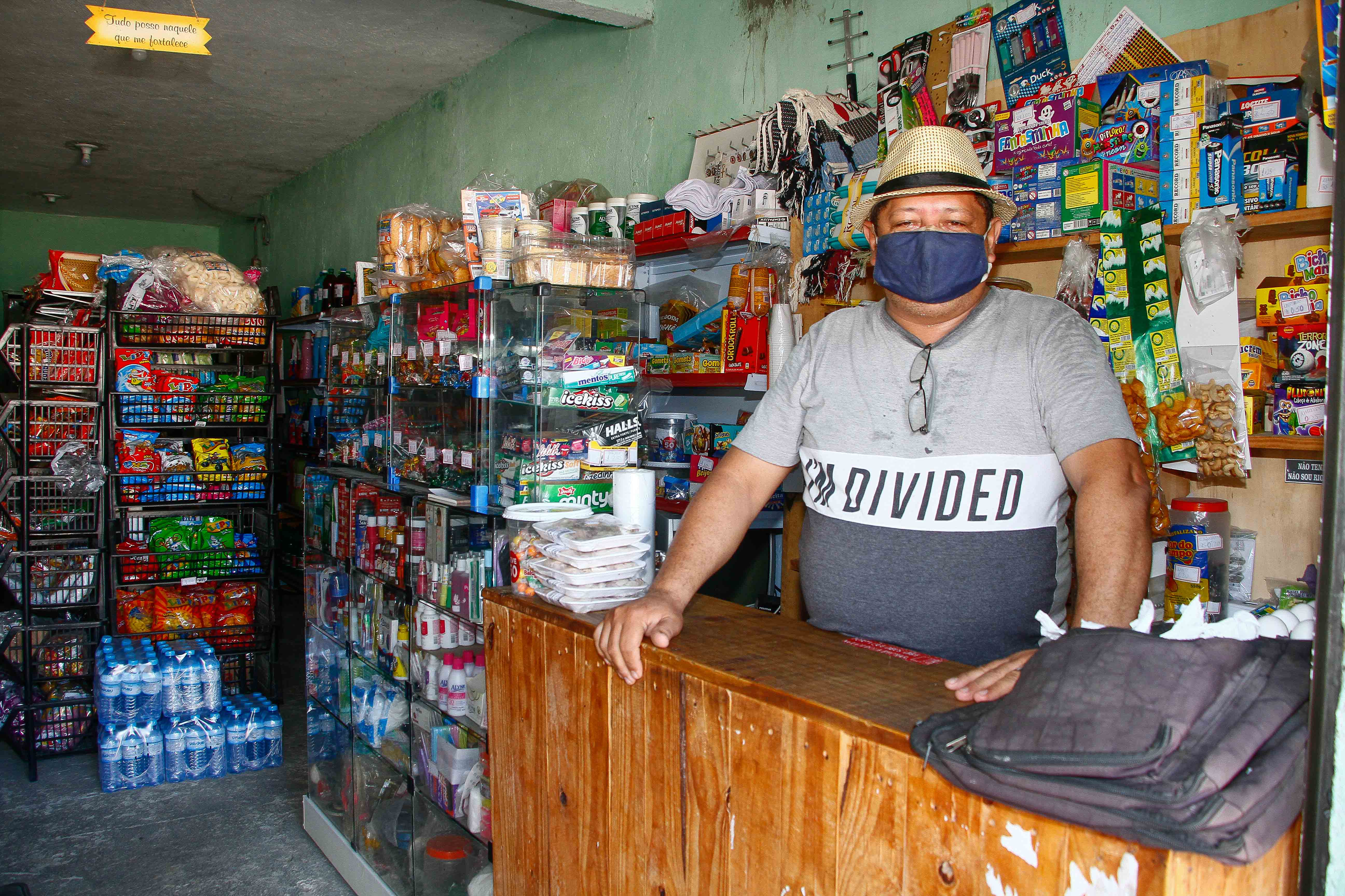 homem de máscara e chapéu posando atrás de balcão de comércio com produtos ao fundo
